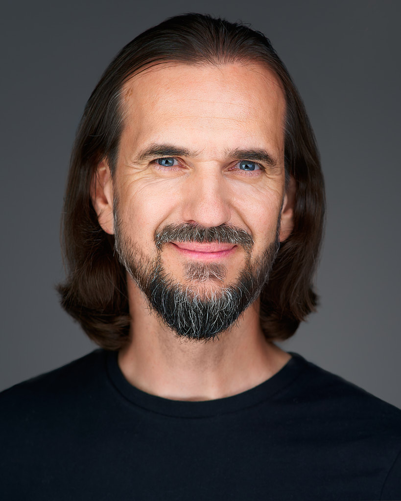 Profesjonalny portret headshot fotografa Marek Wołynko. Portret wykonany na szarym tle. Marek ubrany jest w czarny t-shirt. Ma długie włosy, brodę i przyjazny uśmiech.