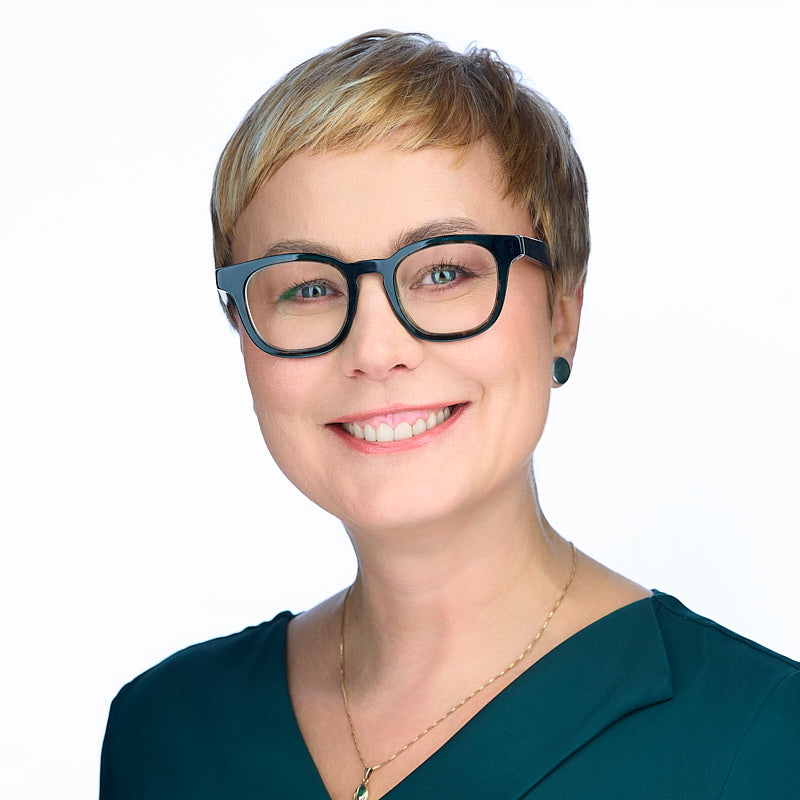 Wysokiej jakości zdjęcie portretowe headshot wykonan podczas sesji dla IBM Polska przez Marka Wołynko. Na zdjęciu pracownica IBM, usmiechnięta. Ubrana w zieloną bluzkę. Na szyi ma łańcuszek. Na oczach świetne okularty z grubymi, ciemnymi oprawkami. Kobieta ma krótkie włosy w kolorze blond. Zdjęcie na białym tle.